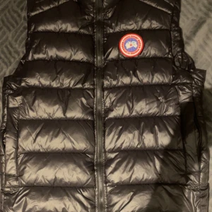 Svart dunväst från Canada Goose - Säljer en svart dunväst från Canada Goose med klassisk logga på bröstet. Västen är quiltad med breda horisontella kanaler och har dragkedja framtill. Perfekt för lager-på-lager och håller dig varm under kyliga dagar. Storlek L passar mig som är runt 180cm. Perfekt för hösten som kommer. Pris kan diskuteras. Använd 1-2 gånger. 