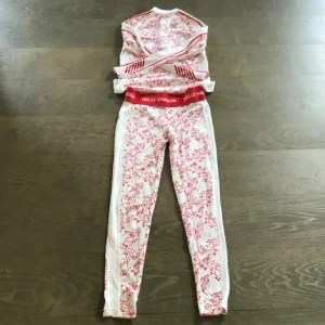 Vit och röd underställströja Helly Hansen - Säljer en vit underställströja från Helly Hansen med halv dragkedja och lång ärm. Tröjan har ett snyggt rött blommigt mönster och logga på kragen. Perfekt för kalla dagar och träning utomhus. Materialet är mjukt och andas, med skön passform.