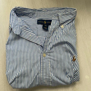 Blåvit randig skjorta från Ralph Lauren - Klassisk skjorta från Ralph Lauren i blåvitt randigt mönster. Skjortan har lång ärm, knappar framtill och broderad logga med polospelare på bröstet. Tillverkad i mjuk bomull och har en snygg krage med knappdetaljer.