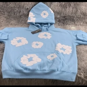 Denim tears hoodie  - Säljer en ljusblå hoodie med stora vita molnliknande mönster över hela tröjan och huvan. Tröjan har huva och en stor magficka framtill. Perfekt för dig som gillar unika och lekfulla plagg.