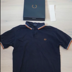 Mörkblå Fred Perry t-shirt S - Snygg mörkblå t-shirt från Fred Perry i 100% bomull. Klassisk pikémodell med korta ärmar och krage med orange och vit randig kant. Ikonisk Fred Perry-logga broderad på bröstet. Perfekt för dig som gillar stilren och sportig vibe.