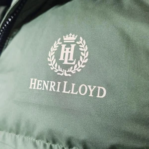 Olivgrön pufferjacka från Henri Lloyd - Snygg olivgrön pufferjacka från Henri Lloyd med broderad logga på bröstet och brittisk flagga på ärmen. Jackan har huva med snörning, dragkedja framtill och stora fickor. Perfekt för kalla dagar och riktigt stilren look.// ser lite off ut i färgen pga ljuset men tveka inte på att att skriva!