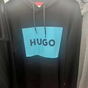 Hugo hoodie - Svart hoodie från HUGO med stor blå rektangel och svart HUGO-logga på bröstet. Tröjan har huva med snörning och en klassisk känguruficka framtill. Perfekt för dig som gillar streetwear och vill sticka ut med en fet logga.