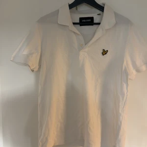 Vit pikétröja från Lyle & Scott - Vit pikétröja från Lyle & Scott med klassisk krage och korta ärmar. Tröjan har den ikoniska gula fågel-loggan broderad på bröstet och knäppning framtill. Perfekt för en clean och stilren look.