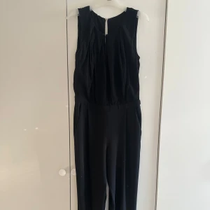 Svart ärmlös jumpsuit - Svart ärmlös jumpsuit med rund hals och snygga veck framtill. Modellen har en markerad midja och raka ben. Stängs med dragkedja i ryggen. Materialet känns mjukt och lätt, perfekt för dig som gillar stilrena plagg