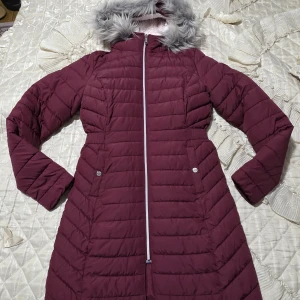 Vinröd lång pufferjacka Hollister - Lång vinröd pufferjacka från Hollister med mysig fuskpäls på huvan och fluffigt vitt foder. Jackan har dragkedja framtill, två fickor med knappar och quiltad design. Perfekt för kalla dagar när du vill hålla stilen och värmen. Köpte för 1500kr från Zalando