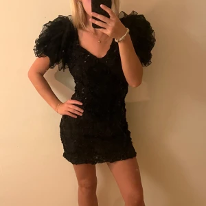 Svart glittrig kortklänning med puffärm - Supersnygg svart kortklänning med stora puffärmar i tyll och glittriga paljetter över hela klänningen. V-ringad och figurnära passform som verkligen sticker ut. Perfekt för dig som vill ha något extra till festen!