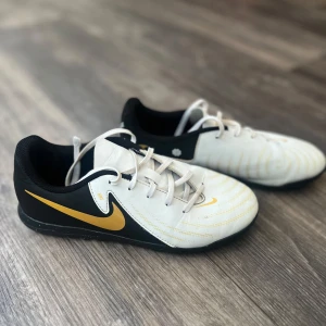 Nike fotbollsskor i vitt och guld - Snygga fotbollsskor från Nike i vitt och svart med guldiga detaljer och swoosh. Skorna har snörning och låg profil, perfekt för snabba rörelser på planen. Ovandelen är i syntetmaterial och sulan är svart. Riktigt stilren design för dig som vill sticka ut på matchen.