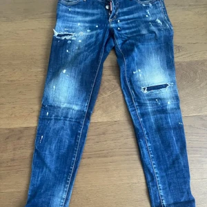 Blå Dsquared2 jeans med slitningar - Snygga blå jeans från Dsquared2 med coola slitningar, färgstänk och broderade detaljer på bakfickorna. Jeansen har en smal passform och klassisk femficksdesign. Tillverkade i bomull med en trendig tvättad look.