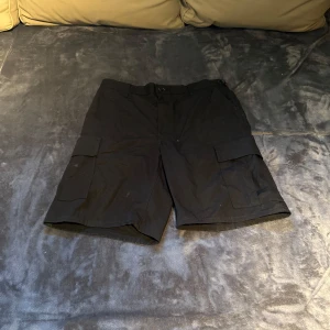 Svarta cargo shorts från Propper - Svarta cargo shorts från Propper i 100% bomull. De har flera stora fickor med lock på sidorna och bak, samt bälteshällor och knappstängning. Perfekta för dig som gillar praktiska och stilrena shorts med loose fit.
