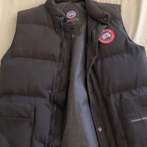 Svart dunväst från Canada Goose - Säljer en svart dunväst från Canada Goose med klassisk logga på bröstet. Västen har två stora fickor framtill, dragkedja och tryckknappar. Perfekt för lager-på-lager. Bara hör av sig vid frågor.😄👍🏽
