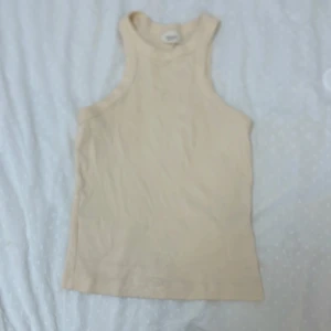 Beige ribbad linnetopp - Säljer en stilren beige linnetopp med hög hals och ärmlös design. Toppen är ribbad och har en tight passform som framhäver axlarna. Perfekt att matcha med jeans eller kjol för en clean look.