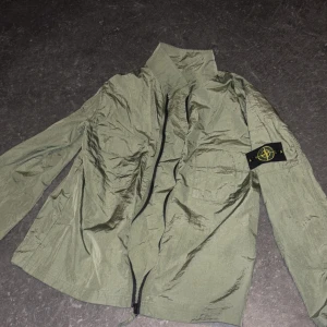 Grön vindjacka från Stone Island - Snygg grön vindjacka från Stone Island med klassisk patch på ärmen. Jackan har hög krage, dragkedja framtill och en bröstficka. Perfekt för dig som gillar streetwear och vill ha en lätt jacka med cool attityd.