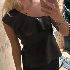 Svart one shoulder topp i skinnimitation - Säljer en svart one shoulder topp med volangdetalj längs halsringningen. Toppen är i ”skinn” material🩷