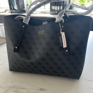  Guess väska - Säljner denna snygga svarta väska från Guess med diskret mönster och guldfärgade detaljer då jag fick dubbletter i present. Väskan är rymlig med långa handtag och inkluderar en matchande mindre väska och plånbok. Nypris ligger på 1695kr. 