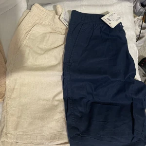 Beige och marinblå linneshorts - Två par shorts från H&M, ett par i beige och ett i marinblått. Båda har elastisk midja med dragsko och sidofickor. Tillverkade i mjukt linnemix som ger en chill och luftig känsla, perfekta för varma dagar. Storlek 170 barn. Både för 320kr 