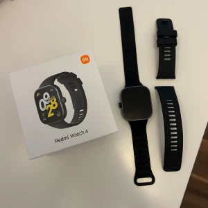Svart Redmi Watch 4 smartklocka - Snygg svart Redmi Watch 4 med fyrkantig display och silikonarmband. Klockan har en modern design med tydlig skärm och medföljande extra armband. Perfekt för dig som gillar teknik och vill ha koll på din vardag direkt på handleden.