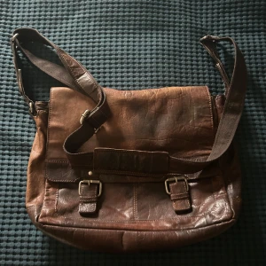 Brun axelväska från Spikes & Sparrow - Snygg brun axelväska i skinn från Spikes & Sparrow med justerbar rem, två spännen framtill och flera praktiska fack. Väskan har ett klassiskt och stilrent utseende med detaljer i metall och randigt innerfoder. Perfekt för dig som vill ha både stil och funktion.