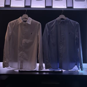 Två Polo Ralph Lauren skjortor slim fit - Två klassiska skjortor från Polo Ralph Lauren i slim fit-modell. En är vit och den andra ljusblå, båda med broderad logga på bröstet och knappar framtill. Perfekt krage och långärmad design, tillverkade i mjuk bomull för en clean och stilren look. Passar mer till kallare tider så bort med linne-skjortorna och in med bomull. Nu säljs två eftertraktade skjortor, skriv om intresse av en eller båda. (Kan förekomma rabatt om du köper båda) storlek XL i barn men passar från 165~175 hör av dig!!