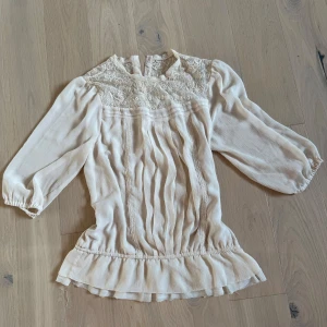 Beige blus med spets och volang - Supersöt beige blus med broderad spets upptill och fina knappar i nacken. Blusen har trekvartsärmar, volangkant nertill och lätt rynkade detaljer framtill. Perfekt för dig som gillar romantisk och vintage-inspirerad stil.