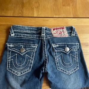 True Religion jeans - y2k - Snygga blå lågmidjade jeans från True Religion med kontrasterande vit söm och ikoniska fickdetaljer med silverknappar. Klassisk femficksmodell och låg midja, även märkespatch baktill. De är ganska raka i modellen men tycker själv de har lite av dn bootcut längst ner.