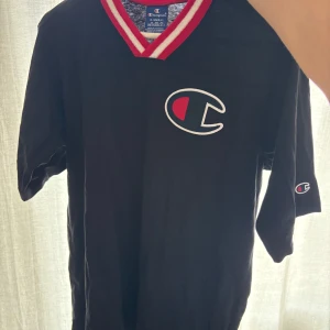 Svart Champion t-shirt med röd/vit krage - Svart t-shirt från Champion i storlek XS med stor broderad logga på bröstet och mindre logga på ärmen. V-ringad krage med röda och vita ränder ger en sportig vibe. Materialet är mjuk bomull och passformen är oversized. Perfekt för dig som gillar streetwear.