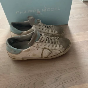 Philippe Model vita sneakers med blå detalj - Snygga sneakers från Philippe Model i vitt skinn med ljusblå detalj på hälen och grå mocka framtill. Klassisk låg modell med snörning och rund tå. Sulan har tydlig Philippe-logga undertill. Perfekta för en avslappnad och trendig look.