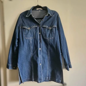 Jeansjacka med nitar från Classic Woman - Snygg jeansjacka i mörkblå denim från Classic Woman. Jackan har långa ärmar, två bröstfickor med coola silverfärgade nitar och knappar framtill. Perfekt oversized look och passar till många olika stilar. fattas 2 knappar men det finns 3 extra knappar på jackan