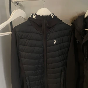 Svart pufferjacka från Peak Performance - Svart pufferjacka från Peak Performance med quiltad framsida och baksida, hel dragkedja och huva med vit logotyptext runt kanten. Jackan har långärmade ärmar och sportig look, perfekt för kyliga dagar.