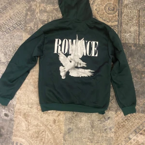Mörkgrön hoodie Romance med tryck - Säljer en mörkgrön hoodie från Romance med stor vit text och duvor tryckta på ryggen. Framsidan har en mindre Romance-logga och en klassisk magficka. Tröjan har huva med snörning och långa ärmar, perfekt för chill dagar.