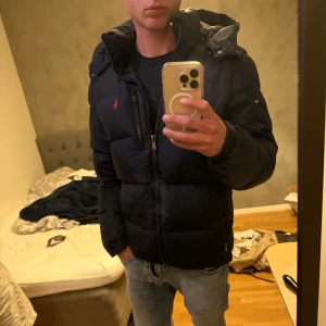 Marinblå dunjacka Polo Ralph Lauren - Säljer en marinblå dunjacka från Polo Ralph Lauren med huva och dragkedja. Jackan har klassisk pufferstil, två sidofickor med dragkedja och ikoniska röda loggan på bröstet. Perfekt för kalla dagar och riktigt snygg till streetwear.