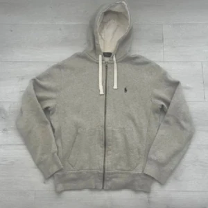 Ralph lauren Hoodie  - Säljer en grå hoodie från Polo Ralph Lauren med vit snörning och dragkedja. Tröjan har huva, två fickor fram och det klassiska Polo-logot broderat på bröstet. Mjuk insida och ribbade muddar gör den extra skön. Perfekt för chill dagar. Nyskick, finns inga tecken på användning. Nypris över 2000:- mitt pris 799:-Storlek S sitter som S/M, frågor? Bara att höra av sig!