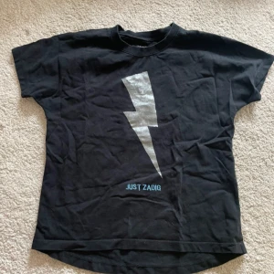 Svart t-shirt med blixt från Zadig - Cool svart t-shirt från Just Zadig med en stor silverfärgad blixt på bröstet. Klassisk rund hals och korta ärmar. Materialet är mjuk bomull och trycket ger en edgy vibe. Perfekt för dig som gillar statement-plagg och vill sticka ut.