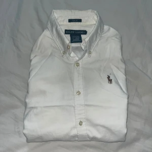 Vit slim fit skjorta från Ralph Lauren - Klassisk vit skjorta från Ralph Lauren i slim fit-modell. Skjortan är tillverkad i 100% bomull och har knappar framtill samt button down-krage. På bröstet finns den ikoniska broderade polospelaren. Perfekt för en clean och stilren look. Storleken är 6/xs-s. Tveka inte att fråga om ni undrar något eller vill ha fler bilder,Mvh Olle 
