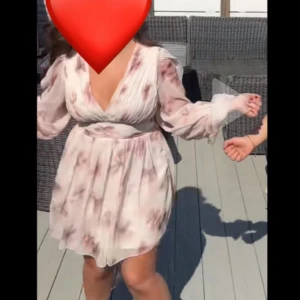 Ljus blommig kortklänning med v-ringning - Säljer en superfin ljus kortklänning med blommigt mönster och v-ringad hals. Klänningen har långa, transparenta ärmar och markerad midja som ger en snygg siluett. Perfekt för dig som gillar romantisk och feminin stil.