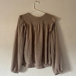 Beige blus med volangdetaljer - Säljer en beige blus med långa ärmar och volangdetaljer vid axlarna. Blusen är tillverkad i ett lätt, luftigt tyg som ger en avslappnad känsla. Perfekt för dig som gillar en romantisk och stilren look.