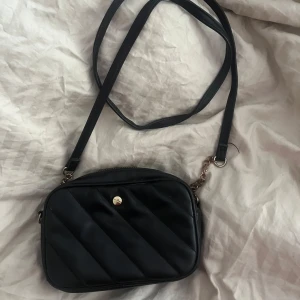 Svart quiltad axelväska med kedjedetalj - Säljer en svart axelväska med quiltad design och guldfärgad kedjedetalj på axelremmen. Väskan har dragkedja upptill och ett litet innerfack. Perfekt storlek för det viktigaste och stilren look.