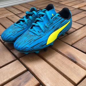 Blå fotbollsskor från Puma - Säljer ett par blå fotbollsskor från Puma med svart och gult mönster. Skorna har snörning och är designade med dobbar för spel på gräs. Perfekta för dig som vill sticka ut på planen!