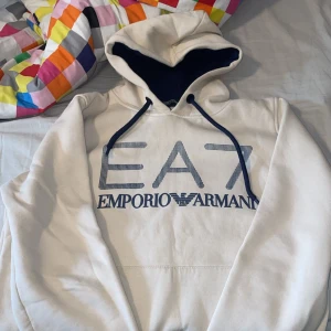 Vit hoodie från Emporio Armani EA7 - Vit hoodie från Emporio Armani EA7 med stor logga framtill. Tröjan har huva med svart innerfoder och svarta snören. Klassisk känguruficka framtill och långa ärmar. Perfekt för en avslappnad och stilren look.
