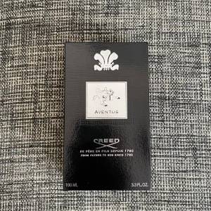Creed Aventus Eau de Parfum 100ml - Creed Aventus är en exklusiv herrparfym i en elegant glasflaska med svart etikett och silverdetaljer. Doften är känd för sina fruktiga och träiga noter. Förpackningen är svart med silverlogga och text. Flaskan rymmer 100 ml.