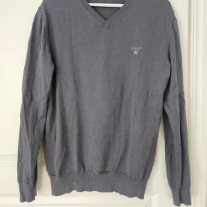 Grå v-ringad tröja från GANT - Säljer en klassisk grå v-ringad tröja från GANT med broderad logga på bröstet. Tröjan har långa ärmar och ribbade muddar vid ärmslut och nederkant. Perfekt för en stilren och avslappnad look.