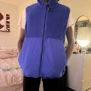 Blå fleeceväst från Adidas - Säljer en blå /lila väst från Adidas med dragkedja framtill. Övre delen är i mjuk fleece och nedre delen i slät polyester. Hög krage och diskret Adidas-logga på bröstet..