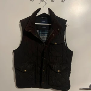 Svart quiltad väst från Gant - Snygg svart quiltad väst från Gant med rutigt innerfoder och bruna detaljer vid kragen. Västen har knappar framtill och två stora fickor med lock. Perfekt för lager-på-lager under kyliga dagar.