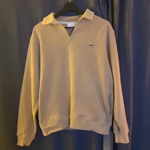 Beige halvzip tröja från Lacoste - Säljer en beige långärmad tröja från Lacoste med halv dragkedja och klassisk krage. Tröjan har det ikoniska gröna krokodilmärket broderat på bröstet och ribbade muddar vid ärmslut och nederkant. Perfekt för en stilren och avslappnad look.