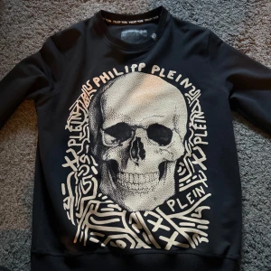 Svart sweatshirt med dödskalle från Philipp Plein - Cool svart sweatshirt från Philipp Plein med stor gul  dödskalle och grafiskt tryck framtill. Tröjan har rund halsringning och är tillverkad i 100% bomull. Perfekt för dig som gillar statement-plagg och streetwear.