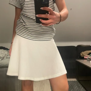 Vit kortkjol i A-linjeform - Säljer en vit kortkjol med A-linjeformad siluett. Kjolen har en enkel och stilren design som passar perfekt till sommaren. Materialet känns lätt och luftigt, vilket gör den bekväm att bära under varma dagar.