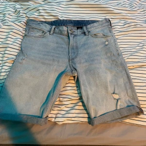 Ljusblå slim fit jeansshorts  - Säljer ett par ljusblå jeansshorts i slim fit-modell. Shortsen har klassisk femficksdesign, uppvikta benslut och slitna detaljer framtill för en trendig look. Perfekta för varma dagar!