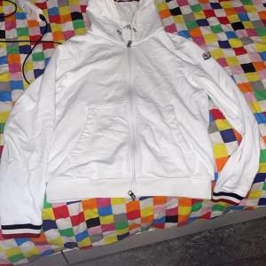 Vit zip-hoodie från Moncler - Säljer en vit hoodie med dragkedja från Moncler. Jackan har huva, två fickor fram och ribbade muddar med röd och marinblå rand. Klassisk Moncler-logga på ärmen och diskreta detaljer på huvan.