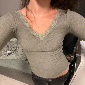 Olivgrön ribbad topp med spetskant - Säljer en olivgrön ribbad långärmad topp med v-ringning och dekorativ spetskant runt halsen. Toppens material är stretchigt och sitter tight mot kroppen. Perfekt för en stilren och enkel look.