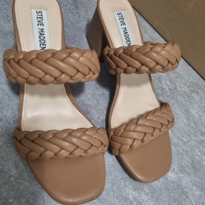 Bruna flätade sandaler från Steve Madden - Snygga bruna sandaler från Steve Madden med två breda flätade remmar och fyrkantig tå. Skorna har en klack och öppen design, perfekta för varmare dagar. Stilrena och trendiga att matcha till många outfits. Dessa har används en gång därför är de smutsiga undersidan, annars är de som nya. Skolådan finns kvar. 